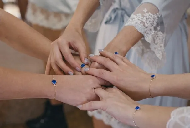 Mãos de noiva e madrinhas sobre renda, com pulseiras de pedra azul